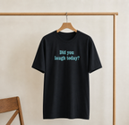 Black t-shirt with 'Did you laugh today?' text on a wooden hanger against a beige wall.