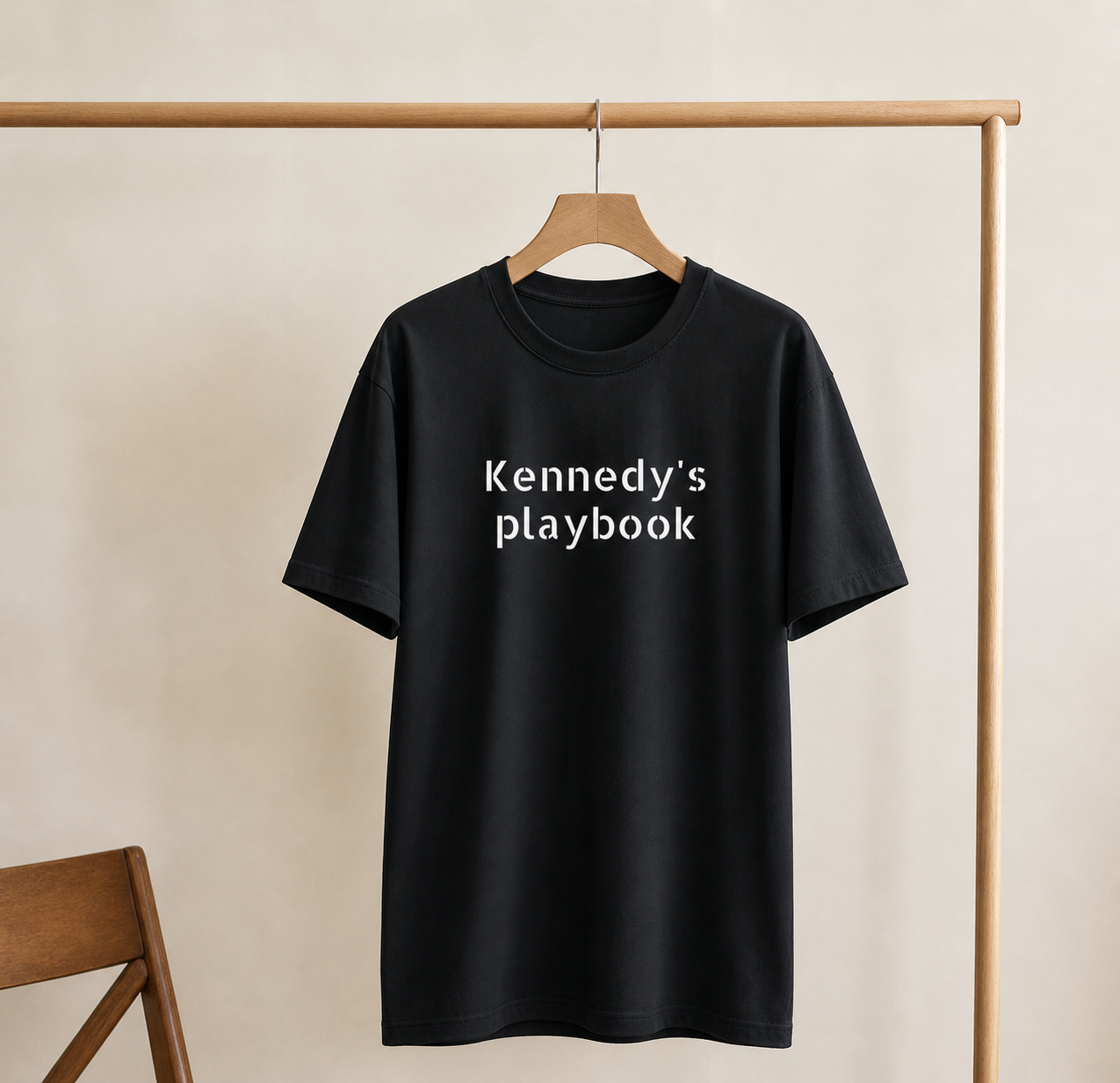 Black t-shirt with 'Kennedy's playbook' text on a wooden hanger against a beige wall.