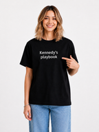 Woman wearing a black t-shirt with 'Kennedy's playbook' text and blue jeans indoors.
