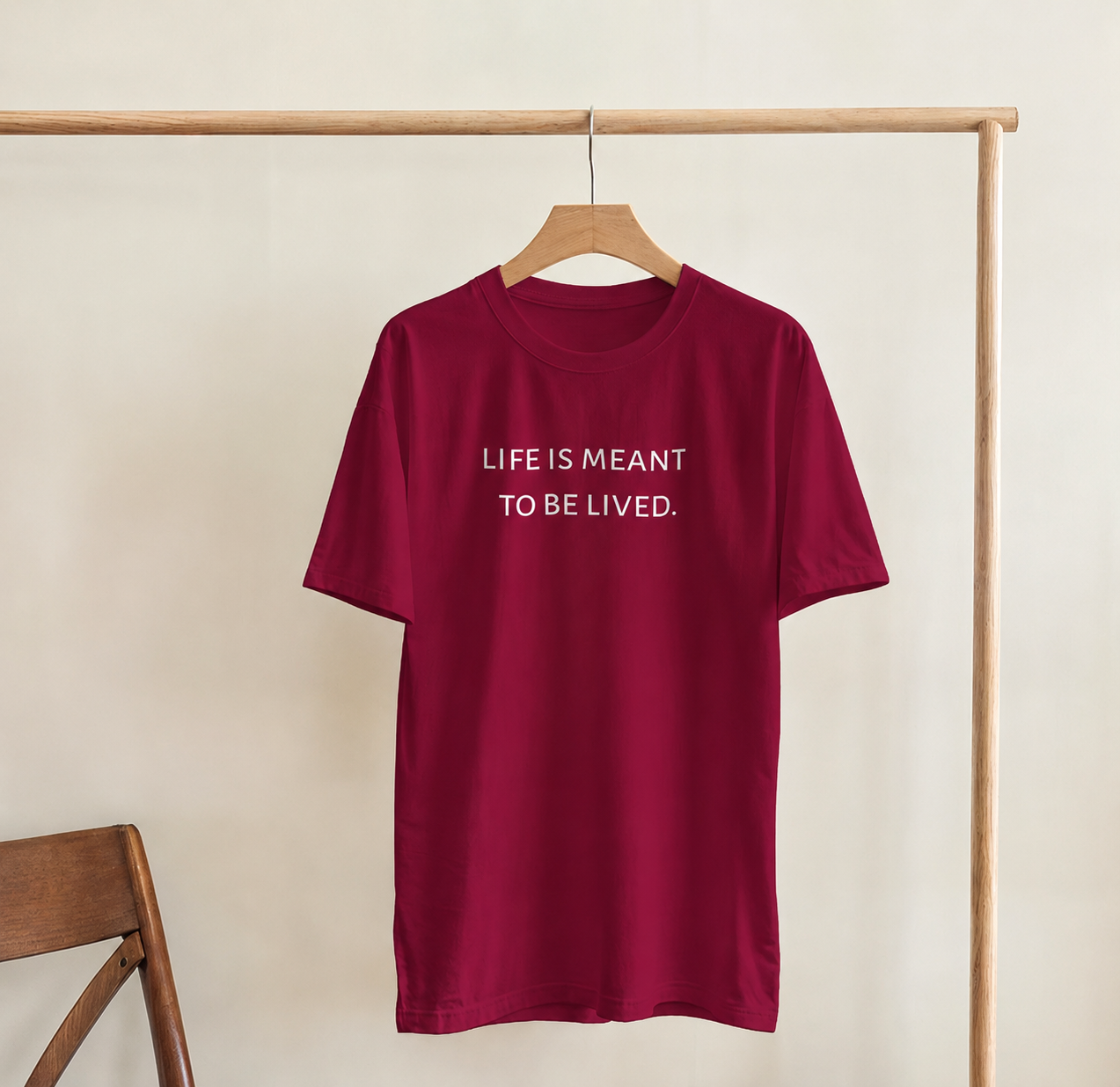 Maroon t-shirt with 'LIFE IS MEANT TO BE LIVED.' text on a wooden hanger against a beige wall.